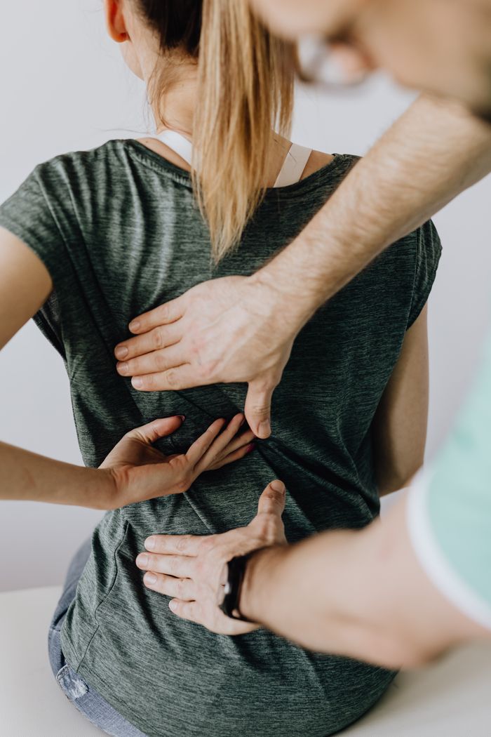 Top view of crop unrecognizable woman in casual wear showing chiropractor in eyeglasses and uniform pain spot on back while sitting on examination couch in hospital